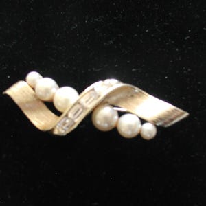 May include: A vintage brooch with a gold-tone metal ribbon design, adorned with faux pearls. The brooch features a series of graduated pearls and a central section with rectangular embellishments. The pin is set against a dark background.