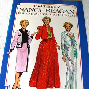 May include: A book titled Tom Tierney Nancy Reagan Fashion Paper Dolls in Full Color featuring illustrations of Nancy Reagan in three different outfits: a pink skirt suit, a red jacket with a long red skirt, and a long white gown with floral patterns.