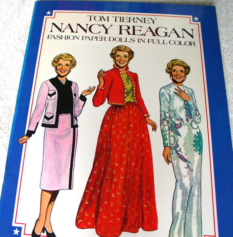 May include: A book titled Tom Tierney Nancy Reagan Fashion Paper Dolls in Full Color featuring illustrations of Nancy Reagan in three different outfits: a pink skirt suit, a red jacket with a long red skirt, and a long white gown with floral patterns.