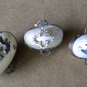 May include: Three oval-shaped, vintage jewelry boxes with silver-toned metal frames. Two boxes feature bird designs, while the center box has a cross-shaped design with a rhinestone border. The boxes have a pearlescent finish.