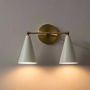 May include: A wall-mounted light fixture with two white, cone-shaped shades. The fixture has a gold-colored circular backplate and a horizontal bar connecting the shades. The light shines downward, illuminating the wall below.