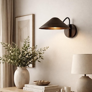 May include: A bronze-colored wall sconce with a conical shade is mounted on a white wall. The sconce has a circular base and a curved arm. A vase of flowers, books, and a table lamp are on a wooden surface.