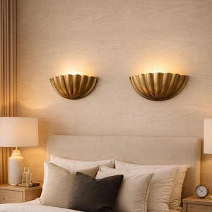 Può includere: Due applique a parete dorate a forma di conchiglia con un bagliore caldo sono montate su una parete con pannelli di legno chiaro. Sotto, un letto è fatto con biancheria da letto e cuscini dai toni neutri. Un comodino con una lampada è visibile.
