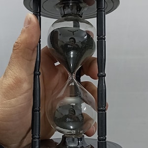 Puede incluir: Un reloj de arena negro con un centro de cristal transparente, lleno de arena negra. El reloj de arena está sostenido por una mano. El marco es de color gris oscuro y tiene una base y una parte superior circulares. Es un artículo decorativo.