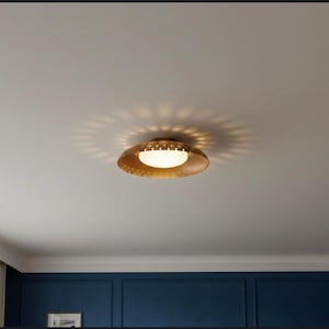 Puede incluir: Una lámpara de techo dorada de estilo moderno de mediados de siglo. La luz tiene una pantalla circular y perforada con un centro de vidrio esmerilado blanco. La lámpara proyecta un patrón de estrella en el techo. La habitación tiene una pared azul y un techo blanco.