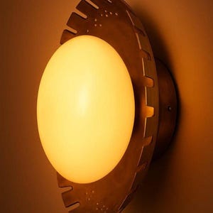 K&ouml;nnte beinhalten: Eine Wandleuchte mit einer warm leuchtenden Kugel, die in einem bronzefarbenen, kreisf&ouml;rmigen Rahmen eingefasst ist. Der Rahmen hat ein einzigartiges, gezahntes Design und ist an einer braunen Wand montiert.