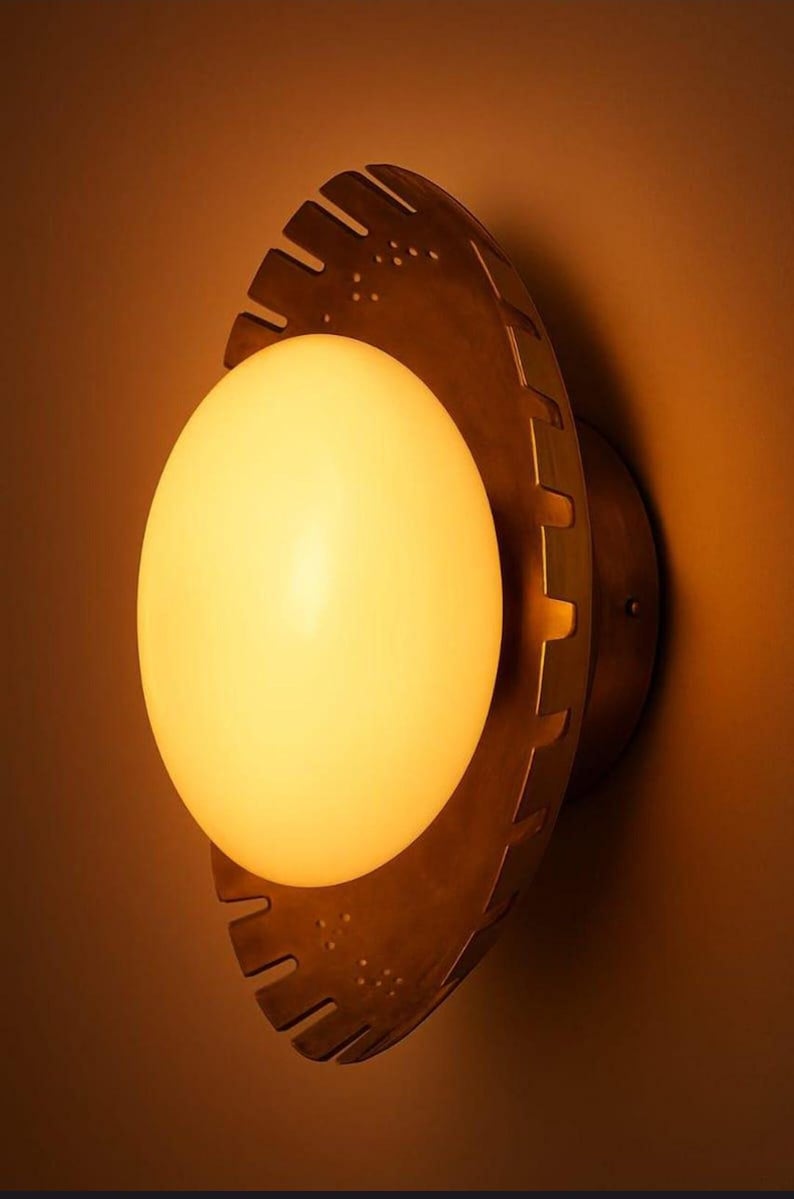 K&ouml;nnte beinhalten: Eine Wandleuchte mit einer warm leuchtenden Kugel, die in einem bronzefarbenen, kreisf&ouml;rmigen Rahmen eingefasst ist. Der Rahmen hat ein einzigartiges, gezahntes Design und ist an einer braunen Wand montiert.