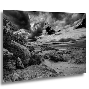 May include: Black and white landscape photograph featuring a dramatic sky filled with dark, swirling clouds. The foreground shows large, textured rocks and sparse vegetation. A small tree stands on a rocky outcrop, with a distant view of a plain.