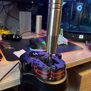 Könnte beinhalten: Ein schwarzer und roter Gaming-Schalthebel mit einem silbernen Metallschaltknüppel. Der Schalthebel ist für die Verwendung mit einem Computer konzipiert und verfügt über eine Reihe von Tasten und Schaltern. Der Hintergrund umfasst einen Computermonitor und anderes Gaming-Zubehör.