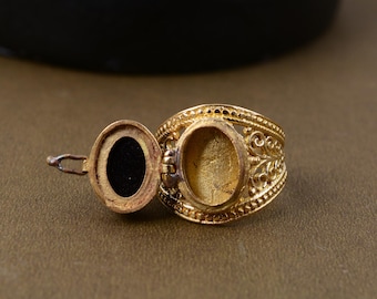 Puede incluir: Un anillo relicario dorado con un compartimento ovalado que se abre para revelar un interior negro. El anillo presenta intrincados detalles de filigrana alrededor de la banda y el marco del relicario. El anillo se muestra sobre una superficie marrón.