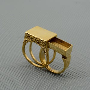 Puede incluir: Un anillo dorado con un compartimento rectangular deslizante. El anillo presenta un diseño ornamental en forma de volutas en los laterales y está montado sobre una doble banda.