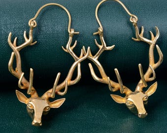 Puede incluir: Un par de pendientes de aro dorados. Cada pendiente presenta una cabeza de ciervo detallada con cornamenta, creando un diseño inspirado en la naturaleza. Los pendientes se muestran sobre un fondo verde oscuro, resaltando su acabado metálico.
