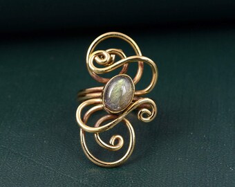 Puede incluir: Un anillo con una piedra preciosa ovalada e iridiscente en el centro, engastada en una estructura de alambre dorado. El alambre metálico forma múltiples bucles y espirales que se extienden verticalmente sobre y bajo la piedra.