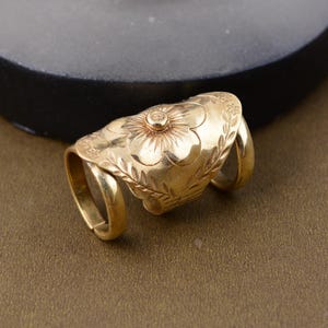 Può includere: Un anello da dito color oro con un design floreale. L'anello presenta un fiore centrale con petali dettagliati ed è circondato da foglie incise. L'anello ha due fasce che si avvolgono attorno al dito.