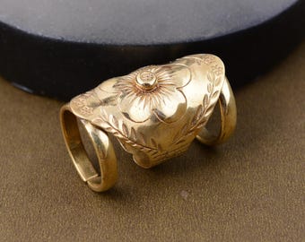 Puede incluir: Un anillo de dedo dorado con un diseño floral. El anillo presenta una flor central con pétalos detallados y está rodeado de hojas grabadas. El anillo tiene dos bandas que se envuelven alrededor del dedo.