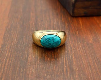 Puede incluir: Un anillo de tono dorado con una banda lisa y pulida, que presenta una piedra turquesa de forma ovalada en el centro. La piedra muestra un color azul vibrante con patrones de vetas oscuras e intrincadas en su superficie, engastada en el metal.