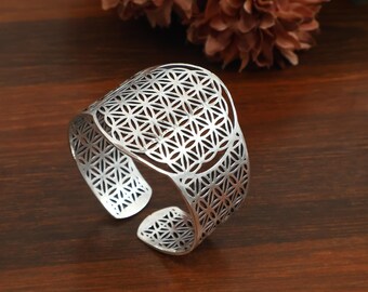 Puede incluir: Un brazalete tipo puño de color plateado que presenta un intrincado patrón geométrico repetitivo de círculos superpuestos, conocido como la flor de la vida. La banda de metal es ancha y abierta, con un diseño calado detallado.