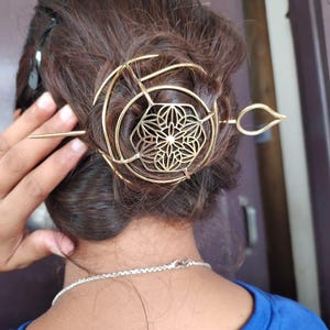 Op de afbeelding: Goudkleurig haaraccessoire met een cirkelvormig ontwerp, een bloemenmiddelpunt en een lange pin. Het haarstuk is geplaatst in donkerbruin haar. Het accessoire is ontworpen om het haar op zijn plaats te houden.