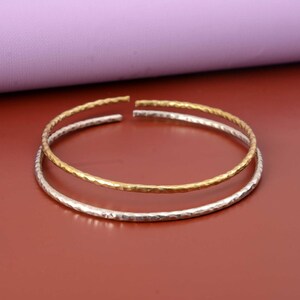 Può includere: Due bracciali sottili, circolari e aperti, uno in tonalità oro lucido e uno in tonalità argento. Entrambi i bracciali presentano una superficie martellata e testurizzata lungo tutta la circonferenza, creando una finitura riflettente.
