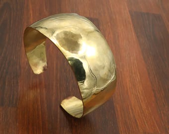 Puede incluir: Una pulsera brazalete ancha de metal dorado pulido con una superficie martillada y texturizada. La pulsera presenta un diseño abierto en la parte posterior con extremos redondeados y un acabado metálico reflectante.