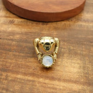 以下が含まれることがあります： 丸い水色の宝石を抱えた金色のカエルリング。リングは光沢のある金属製で、木製の表面に置かれています。カエルのデザインは詳細で、足や質感のある体が見えます。