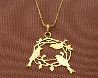 Puede incluir: Un collar de tono dorado con un colgante circular que presenta un diseño recortado de tres pájaros posados en un nido de ramas y hojas entrelazadas. El colgante cuelga de una cadena fina de tono dorado a juego con un acabado liso y pulido.