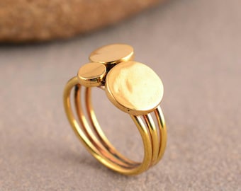 Puede incluir: Un anillo de tono dorado con tres bandas finas apiladas que convergen en un solo cuerpo. La parte superior del anillo está adornada con tres discos circulares planos de diferentes tamaños, dispuestos en un grupo con un acabado metálico pulido.