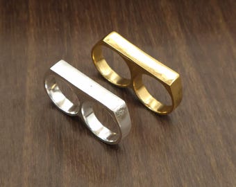 Puede incluir: Dos anillos de doble dedo, uno con acabado plateado pulido y otro con acabado dorado pulido. Cada anillo presenta dos aberturas circulares conectadas por una barra rectangular lisa y única que se extiende sobre la parte superior de los dedos.
