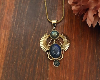 Puede incluir: Un collar de tono dorado con un colgante que presenta tres piedras ovaladas de color azul oscuro. La piedra central es la más grande, flanqueada por dos piedras más pequeñas. El engaste metálico incluye estructuras aladas simétricas y una cadena fina.