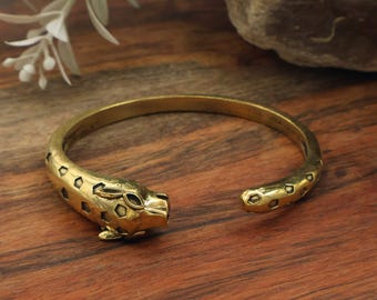 Puede incluir: Un brazalete de metal en tono dorado con una cabeza de animal esculpida en un extremo. La banda está decorada con un patrón repetitivo de formas hexagonales hundidas que se extienden a lo largo de la estructura circular abierta.