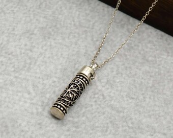 Puede incluir: Collar con colgante plateado. El colgante cilíndrico presenta un diseño intrincado con un patrón similar a un árbol. El collar tiene una cadena delicada y elementos decorativos en la parte superior e inferior.