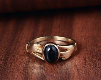 Puede incluir: Un anillo dorado con una piedra negra ovalada. El anillo tiene un diseño único con elementos estilizados en forma de mano a ambos lados de la piedra. Se encuentra sobre una superficie de madera marrón.