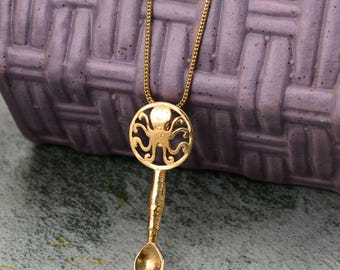 Puede incluir: Un collar de tono dorado con un colgante en forma de cuchara pequeña con una parte superior circular. La sección circular muestra un diseño recortado de un pulpo con tentáculos curvos. El colgante cuelga de una cadena fina de metal dorado.