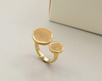 Puede incluir: Un anillo de tono dorado con un diseño de banda abierta que presenta dos discos circulares lisos y planos de diferentes tamaños. El metal tiene un acabado pulido y reflectante, y la estructura es continua, uniendo ambos elementos.