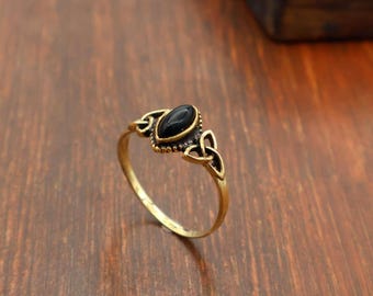 Puede incluir: Un anillo de metal en tono dorado con una piedra negra central en forma de marquesa. El engaste presenta detalles intrincados de nudos a ambos lados de la piedra, con una banda fina y lisa que completa el diseño de esta joya.