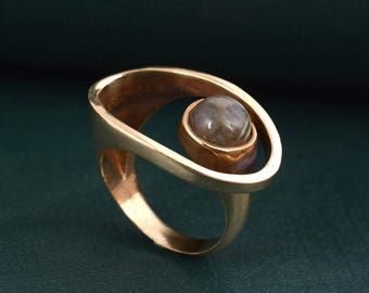 Puede incluir: Un anillo de tono dorado con un diseño de banda ovalada abierta y asimétrica. Una piedra única, lisa, redonda y de color gris parduzco está engastada en un bisel circular unido a un lado de la banda abierta, creando un efecto de espacio negativo.