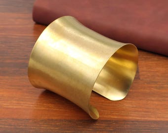 Puede incluir: Un brazalete ancho de color dorado con un diseño curvo. El brazalete está hecho de un metal brillante y se encuentra sobre una superficie de madera. Un cuaderno de cuero rojo oscuro está en el fondo.