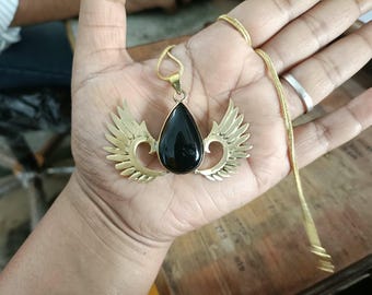 Puede incluir: Un collar de tono dorado con un colgante central de piedra negra en forma de lágrima. El colgante está flanqueado por dos alas metálicas simétricas de tono dorado que se extienden hacia afuera. La pieza cuelga de una cadena fina a juego.