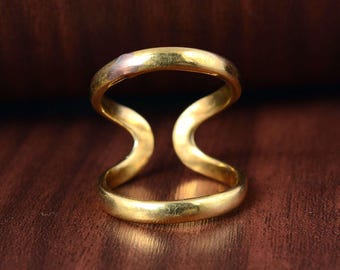 Puede incluir: Un anillo dorado con un diseño único de doble banda. El anillo presenta una forma continua y fluida con dos bandas curvas conectadas por un espacio central abierto. El anillo se muestra sobre una superficie marrón.