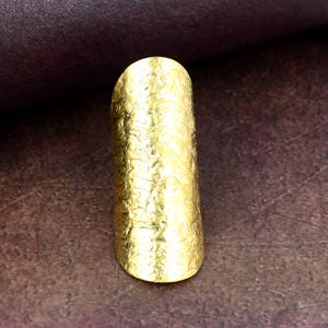 Könnte beinhalten: Ein goldfarbener Ring mit strukturierter Oberfläche. Der Ring hat eine längliche, ovale Form und besteht aus einem glänzenden, metallischen Material. Der Ring ist ein Schmuckstück.