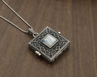 Puede incluir: Un colgante de relicario cuadrado de plata en una cadena fina, con un intrincado patrón en relieve con volutas y formas de diamante. Una piedra lunar iridiscente, lisa y de corte cuadrado, está engastada en el centro del relicario con bisagra.