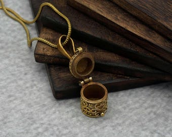 Puede incluir: Un colgante de medallón dorado abierto con un diseño intrincado. El colgante está en una cadena dorada y tiene un compartimento redondo con bisagras. La parte inferior del medallón presenta un patrón calado detallado. El fondo es una superficie de madera oscura.
