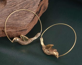 Puede incluir: Un par de pendientes de aro dorados con un dije de pájaro texturizado unido a cada alambre circular fino. Los pájaros tienen marcas detalladas en las alas y el cuerpo con un acabado metálico, y los aros cuelgan del pico y la cola del ave.