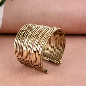 May include: A wide, gold-colored cuff bracelet. The bracelet is made of multiple thin, textured bands that curve around the wrist. The metal has a slightly aged appearance, giving it a vintage feel. The cuff is a statement piece of jewelry.