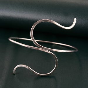 May include: A silver-toned arm cuff with a unique, flowing design. The cuff features a slender band that wraps around the arm, with two curved, open-ended sections that create an elegant, abstract shape. The metal has a polished finish.