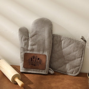 May include: A striped oven mitt and pot holder set with a brown leather patch that reads "MOM FLAVOR" with kitchen utensils. A wooden rolling pin is next to the set. The items are brown and white.