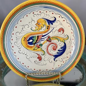 May include: A decorative ceramic plate with a yellow, green, and blue rim. The plate features a central design of a stylized, colorful creature with a yellow head and blue and green wings, surrounded by intricate scrollwork.