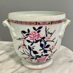 May include: A white ceramic planter featuring a hand-painted floral design with pink blossoms and dark blue leaves. The vessel has two side handles, a flared rim with a red geometric border, and a pedestal base. The exterior is decorated with branches.