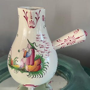 May include: Cream-colored ceramic pitcher with a bulbous body and a handle. The pitcher features a painted scene of a person playing a wind instrument, with red and green floral accents. The pitcher has three small feet and a decorative rim.