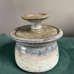 May include: Handmade ceramic butter dish with a lid and a pedestal handle. The dish features a speckled white base, transitioning to a pink and blue band, and topped with a tan lid and handle. The pottery has a rustic, artisanal aesthetic.